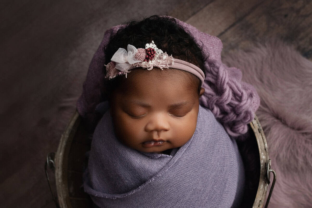 A sleeping baby wrapped in a soft purple blanket, wearing a decorative headband with flowers and berries, lies in a small basket on a wooden floor with a fluffy purple rug nearby. Melinda Gilmore Photography - Maternity & Newborn Photographer - Louisiana