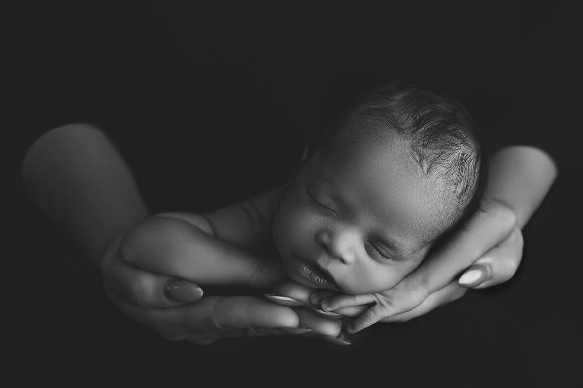 A newborn baby peacefully sleeps with their head resting in gently cupped adult hands, set against a dark background. The image is black and white, highlighting the serene and protective atmosphere. Melinda Gilmore Photography - Maternity & Newborn Photographer - Louisiana