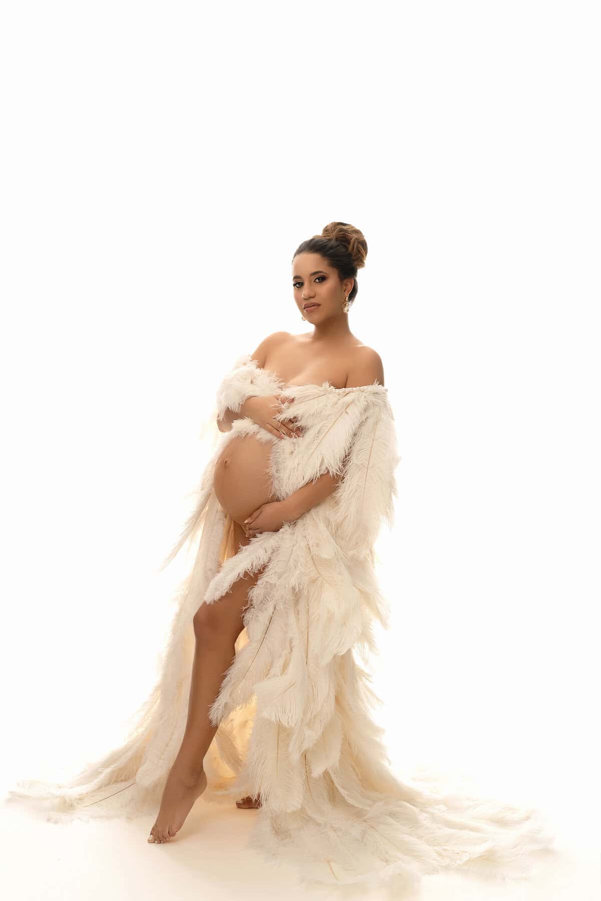 A pregnant woman stands barefoot against a white background, wearing a long, flowing, off-shoulder gown with feather-like textures. She holds her belly with both hands and looks confidently at the camera. Melinda Gilmore Photography - Maternity & Newborn Photographer - Louisiana