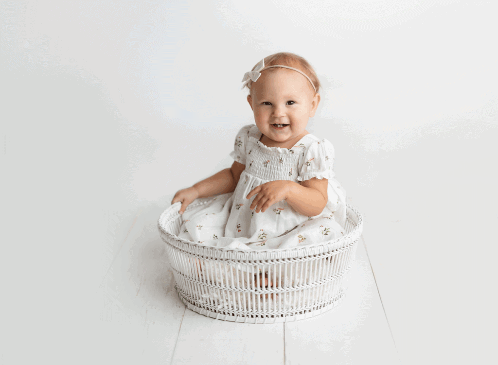 A smiling baby wearing a white dress with a floral pattern and a bow headband sits inside a white wicker basket on a white wooden floor against a plain white background. Melinda Gilmore Photography - Maternity & Newborn Photographer - Louisiana