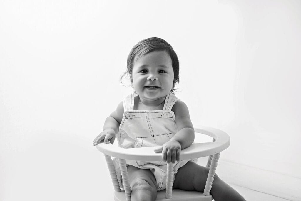 Black and white photo of a smiling toddler with dark hair sitting in a small wooden chair, wearing light-colored overalls, against a plain, bright background. Melinda Gilmore Photography - Maternity & Newborn Photographer - Louisiana