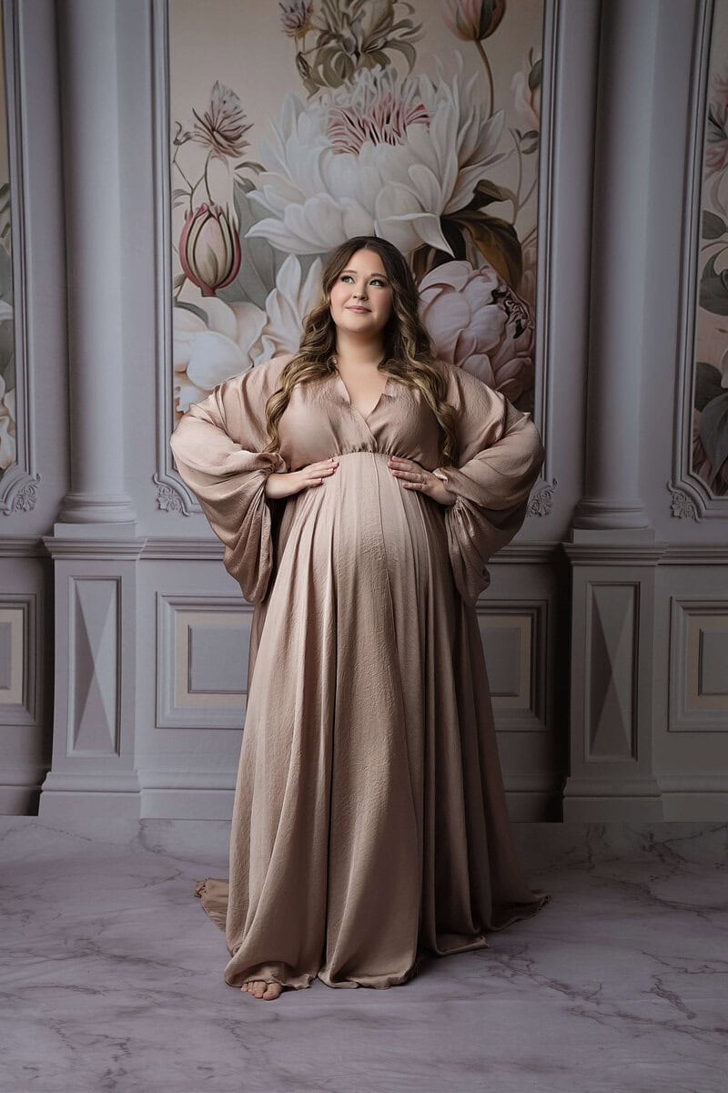 A pregnant woman in a flowing beige gown stands barefoot in front of a decorative floral backdrop, gently cradling her belly and looking upward with a serene expression. Melinda Gilmore Photography - Maternity & Newborn Photographer - Louisiana