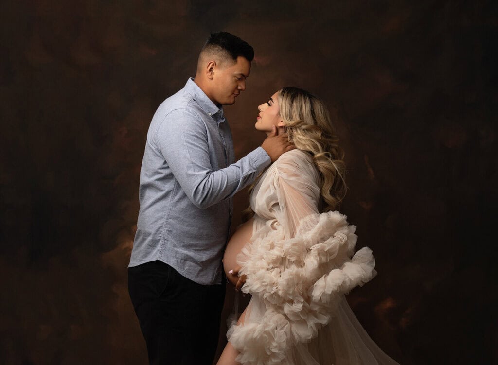 A man gently holds a pregnant woman's chin as they gaze at each other. The woman wears a sheer, ruffled gown and rests her hand on her belly, while the man wears a light blue shirt and black pants. The background is dark and blended. Melinda Gilmore Photography - Maternity & <a href=