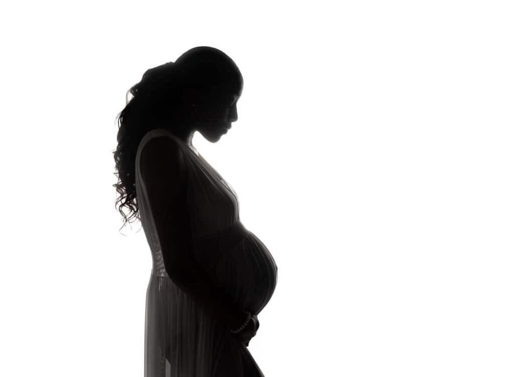 Silhouette of a pregnant woman in profile, gently holding her belly, wearing a flowing dress, with her long hair cascading over her shoulder against a white background. Melinda Gilmore Photography - Maternity & Newborn Photographer - Louisiana