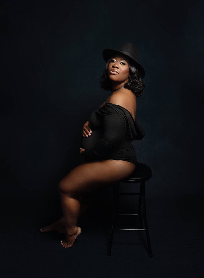 A pregnant woman in a black off-shoulder bodysuit and black hat sits barefoot on a stool against a dark background, cradling her belly and looking confidently at the camera. Melinda Gilmore Photography - Maternity & Newborn Photographer - Louisiana