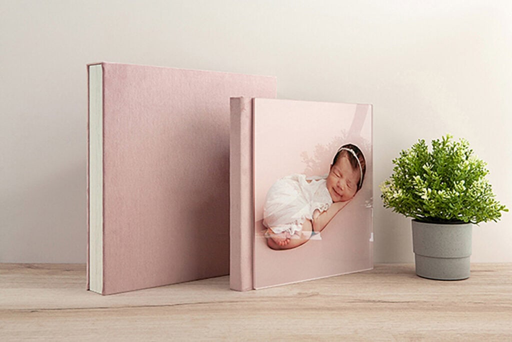 Two pink photo albums are displayed on a wooden surface; one features a cover with a sleeping baby in white clothes. A small green potted plant sits beside the albums against a light neutral wall. Melinda Gilmore Photography - Maternity & Newborn Photographer - Louisiana