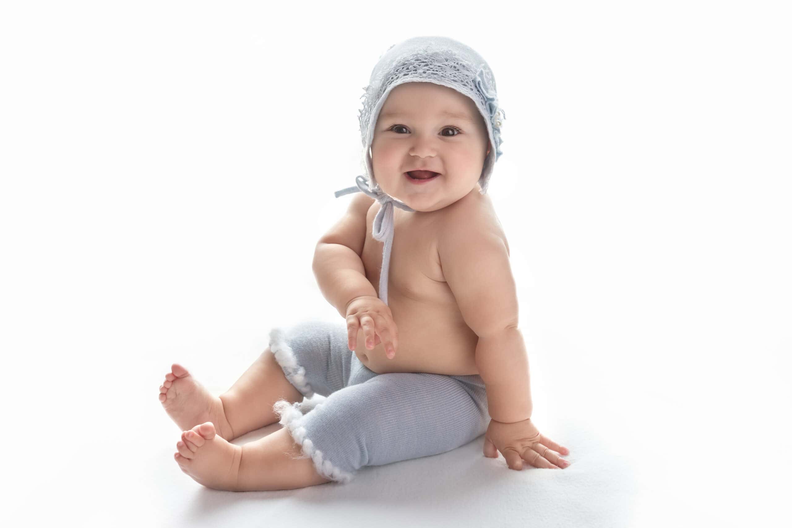 Smiling baby boy sitting on white backdrop wearing a soft knit bonnet and neutral pants during milestone photography session in Morgan City Louisiana
